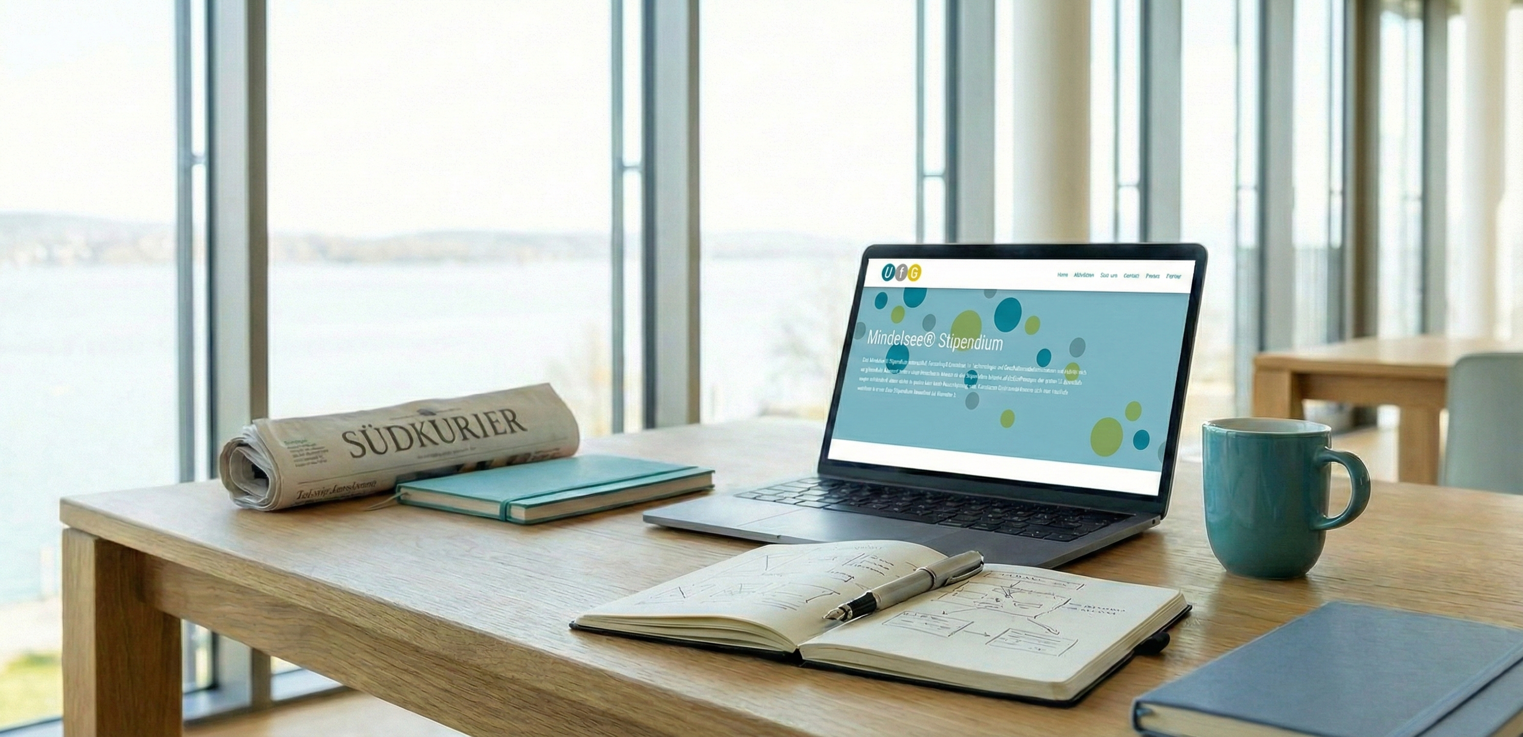 Moderner Arbeitsplatz vor hellen Fenstern mit Seeblick, darauf ein Laptop mit der Mindelsee-Stipendium Website, Notizbücher und eine eingerollte Südkurier-Zeitung